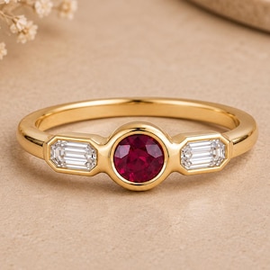 Pode incluir: Um anel de ouro com um rubi vermelho central de corte redondo em engaste de bisel, ladeado por dois diamantes brancos de corte baguette em cada lado. A banda é lisa e polida, apresentando um design clássico de três pedras centrais.