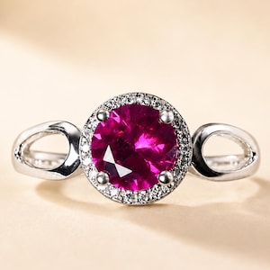 Könnte beinhalten: Ein silberfarbener Ring mit einem großen, runden, leuchtend magentafarbenen Mittelstein. Der Mittelstein ist von einem Kranz aus kleinen, klaren, funkelnden Steinen umgeben. Die Ringschiene ist beidseitig gespalten und bildet eine offene Schlaufe.