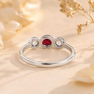 May include: A silver-toned ring featuring a central round-cut red gemstone flanked by two smaller round-cut clear white gemstones. The stones are set in a bezel style along a smooth, polished metal band with a rounded profile and a consistent width.