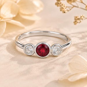 May include: A silver-toned ring featuring a central round-cut deep red gemstone flanked by two smaller round-cut clear white gemstones. Each stone is set in a smooth bezel setting on a polished metal band with a rounded profile and high shine.