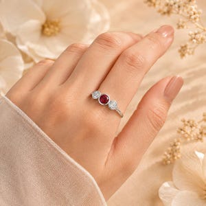 May include: A silver-toned ring featuring a central round-cut red gemstone flanked by two smaller round-cut clear white gemstones, all set in a bezel style on a thin, polished metal band that encircles the finger with a sleek, minimalist design.