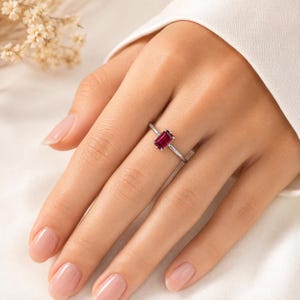 Op de afbeelding: Een zilverkleurige ring met een enkele, smaragdgeslepen, dieprode edelsteen in een zetting met vier pootjes. De ringband is dun en gepolijst, met een strak ontwerp dat de rechthoekige vorm en de levendige kleur van de steen benadrukt.