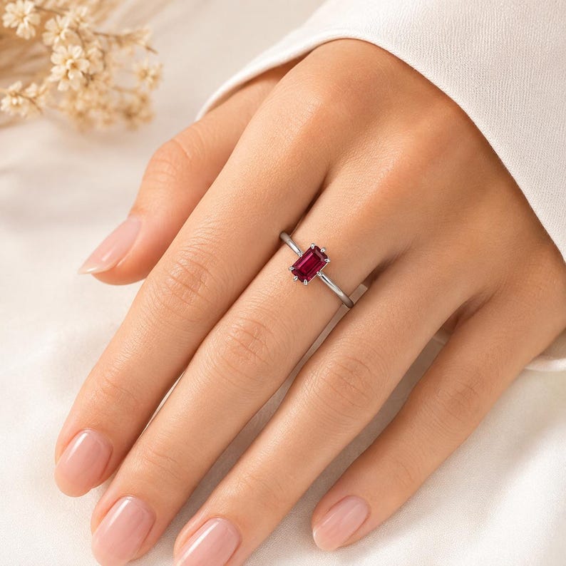 Op de afbeelding: Een zilverkleurige ring met een enkele, smaragdgeslepen, dieprode edelsteen in een zetting met vier pootjes. De ringband is dun en gepolijst, met een strak ontwerp dat de rechthoekige vorm en de levendige kleur van de steen benadrukt.