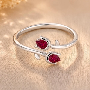 May include: A silver bypass ring featuring two tulip-shaped motifs, each set with a pear-cut, deep red gemstone. The band is polished silver, and the design shows two flower buds facing opposite directions, creating an open, adjustable-style ring.