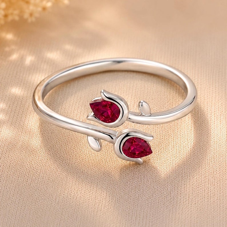 May include: A silver bypass ring featuring two tulip-shaped motifs, each set with a pear-cut, deep red gemstone. The band is polished silver, and the design shows two flower buds facing opposite directions, creating an open, adjustable-style ring.