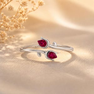 May include: A silver-toned open ring featuring two pear-shaped, deep red stones set in tulip-shaped metal casings. The band is slender and polished, with the two floral-inspired ends overlapping slightly to create a bypass-style design.