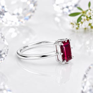 Op de afbeelding: Een zilverkleurige ring met een rechthoekige, smaragdgeslepen dieprode edelsteen in een zetting met vier pootjes. De ringband is glad en gepolijst, wat zorgt voor een strak ontwerp dat de levendige kleur en helderheid van de steen benadrukt.