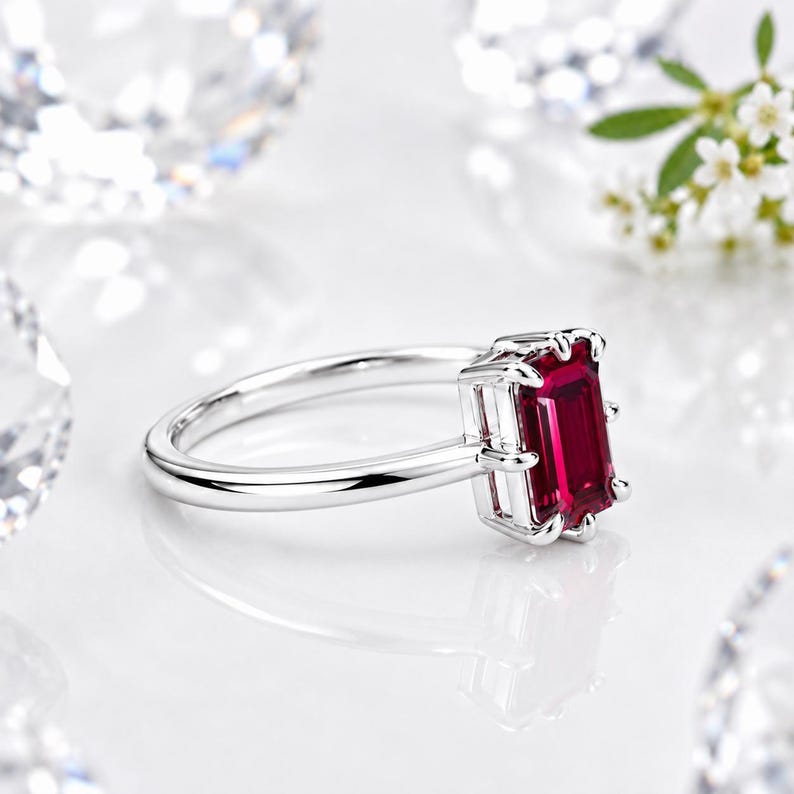 Op de afbeelding: Een zilverkleurige ring met een rechthoekige, smaragdgeslepen dieprode edelsteen in een zetting met vier pootjes. De ringband is glad en gepolijst, wat zorgt voor een strak ontwerp dat de levendige kleur en helderheid van de steen benadrukt.
