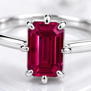 Op de afbeelding: Een zilverkleurige ring met een opvallende rechthoekige, dieprode edelsteen in emerald-slijpvorm, gezet in een zetting met zes pootjes. De ringband is glad en gepolijst en heeft een strakke, reflecterende afwerking die de rode steen accentueert.