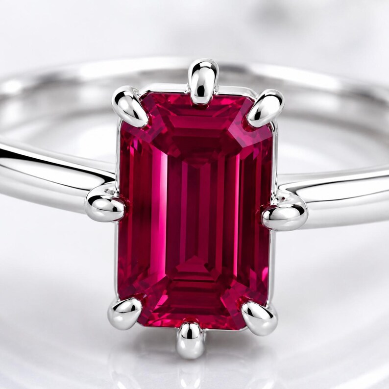 Op de afbeelding: Een zilverkleurige ring met een opvallende rechthoekige, dieprode edelsteen in emerald-slijpvorm, gezet in een zetting met zes pootjes. De ringband is glad en gepolijst en heeft een strakke, reflecterende afwerking die de rode steen accentueert.