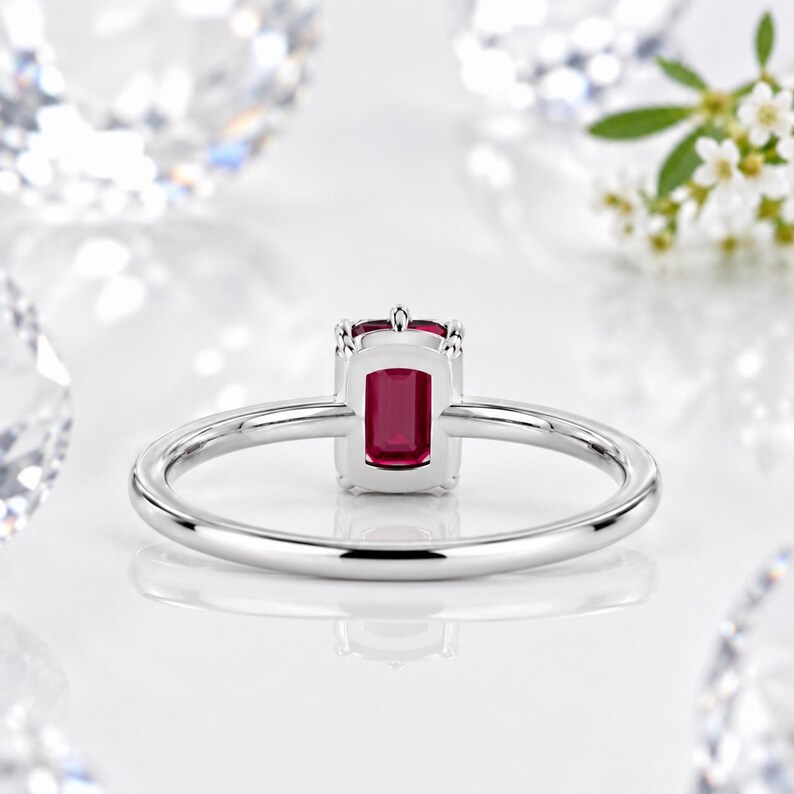 Op de afbeelding: Een zilverkleurige ring met een rechthoekige, dieprode edelsteen in een zetting met vier pootjes. De ringband is glad en gepolijst en heeft een strak, eenvoudig ontwerp dat de levendige kleur van de centrale steen benadrukt.