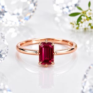 Op de afbeelding: Een ring van ros&eacute;goud met een centrale, smaragdgeslepen, dieproze edelsteen die door zes pootjes wordt vastgehouden. De ringband is glad en gepolijst en toont een strak ontwerp dat de levendige kleur en rechthoekige vorm benadrukt.