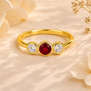 May include: A gold ring featuring a central round-cut red gemstone flanked by two smaller round-cut clear white gemstones. Each stone is set in a smooth bezel setting along the thin, polished gold band, creating a balanced three-stone design.