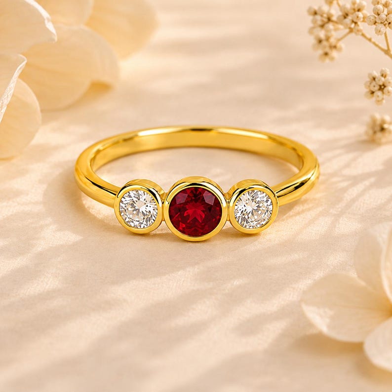May include: A gold ring featuring a central round-cut red gemstone flanked by two smaller round-cut clear white gemstones. Each stone is set in a smooth bezel setting along the thin, polished gold band, creating a balanced three-stone design.