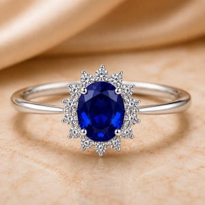 Könnte beinhalten: Ein silberfarbener Ring mit einem zentralen, oval geschliffenen, tiefblauen Edelstein, der von einem Kranz aus kleinen, runden, klaren weißen Steinen umgeben ist. Die Ringschiene ist schmal und poliert und bildet einen klassischen Look.