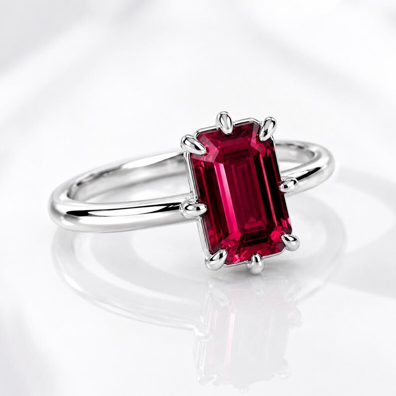 Op de afbeelding: Een zilverkleurige ring met een rechthoekige, smaragdgeslepen dieprode edelsteen in een klauwzetting. De ringband is glad en gepolijst en vertoont een strakke metalen afwerking die de levendige, verzadigde kleur van de centrale steen aanvult.