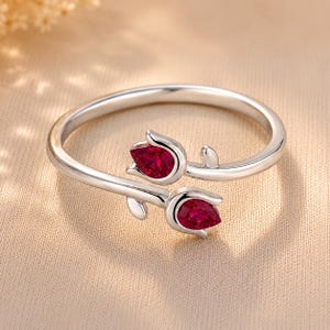 May include: A silver bypass ring featuring two tulip-shaped motifs, each set with a pear-cut, deep red gemstone. The band is polished silver, and the design shows two flower buds facing opposite directions, creating an open, adjustable-style ring.