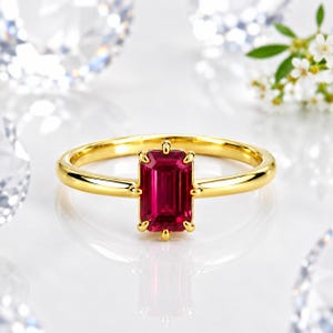 Op de afbeelding: Een gouden ring met een enkele, smaragdgeslepen, levendig rode edelsteen in een zetting met vier pootjes. De ringband is glad en gepolijst en toont een strak ontwerp dat de diepe karmozijnrode kleur en rechthoekige vorm van de steen benadrukt.