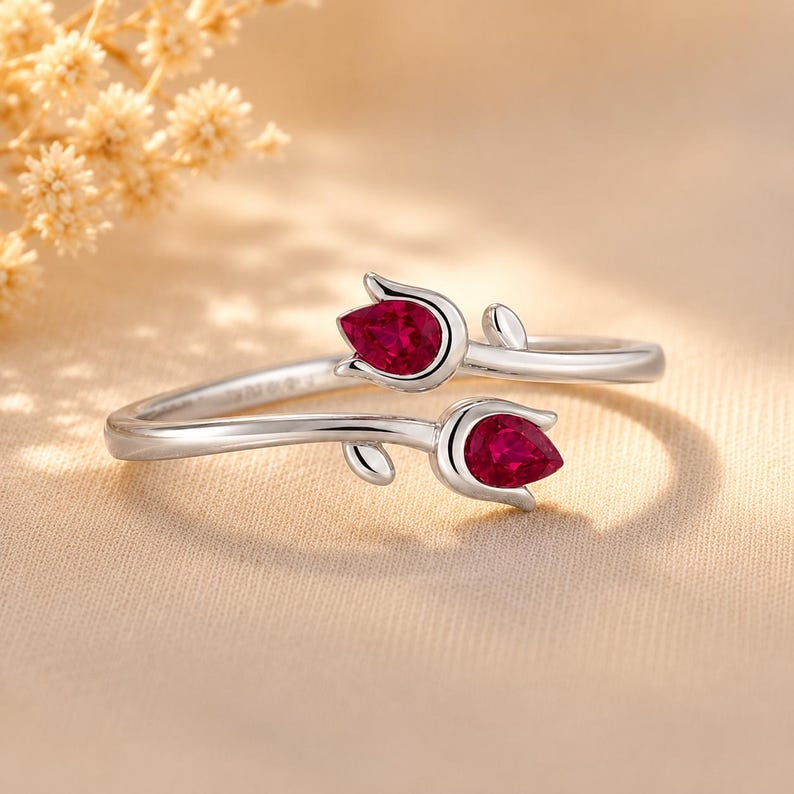 May include: A silver bypass ring featuring two tulip-shaped settings, each holding a pear-cut vibrant red gemstone. The metal band is polished and smooth, with the two floral ends overlapping slightly to create an open, adjustable design.