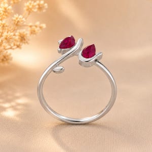 May include: A silver-toned open ring featuring two teardrop-shaped deep red stones set at the ends of the band. The band has a smooth, polished finish and includes a small leaf-like detail on one side near the stone setting.