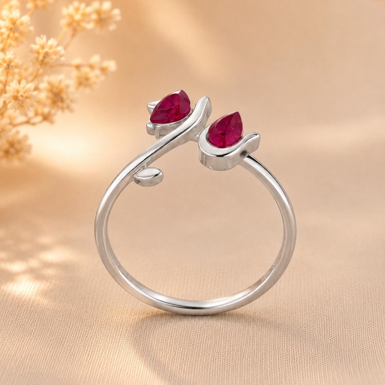 May include: A silver-toned open ring featuring two teardrop-shaped deep red stones set at the ends of the band. The band has a smooth, polished finish and includes a small leaf-like detail on one side near the stone setting.