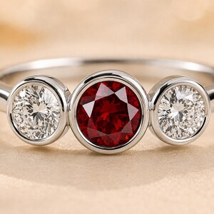 May include: A silver-toned ring featuring a three-stone design with a central round-cut deep red gemstone flanked by two smaller round-cut clear white gemstones. Each stone is secured in a smooth bezel setting on a polished metal band.