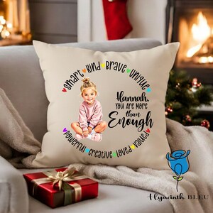 May include: A cream cushion with a print of a young girl in a pink sweater. The text reads "Hannah, You Are More Than Enough" with words like "smart", "kind", and "brave" around the girl. A red gift box with a gold ribbon is in the foreground.