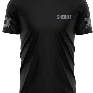 May include: Black short-sleeved t-shirt with the word "SHERIFF" printed on the front. The sleeves have a faded American flag design.