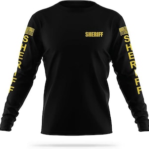 May include: Black long-sleeve shirt with yellow text that reads "SHERIFF" on the front and "SHERIFF" vertically down each sleeve.  The sleeves also feature a small American flag graphic.