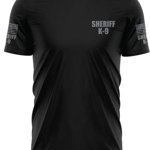 May include: Black crew neck t-shirt with the words "SHERIFF K-9" printed in gray on the chest. The sleeves feature the American flag and the words "SHERIFF K-9" printed in gray. The shirt is made of a smooth, likely athletic material.