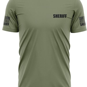 May include: A green short-sleeved t-shirt with the word "SHERIFF" printed on the front. The shirt has an American flag design on each sleeve.