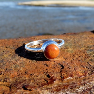 May include: A silver ring featuring a round, orange agate gemstone. The ring is displayed on a rusty brown surface.