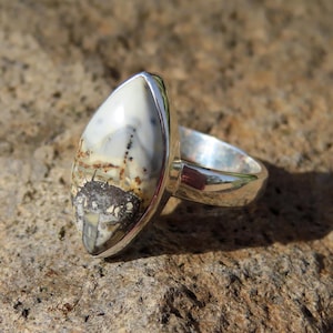 May include: A silver ring with a large, oval gemstone displaying a distinctive pattern of white, grey, and brown. The stone is set in a polished silver band. The ring is presented on a textured, neutral-toned surface. The ring's design is eye-catching.
