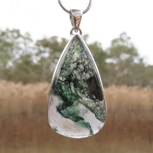 May include: A teardrop-shaped pendant with a silver setting. The pendant has a translucent stone with green, black, and white inclusions, like a landscape. It hangs from a silver chain.