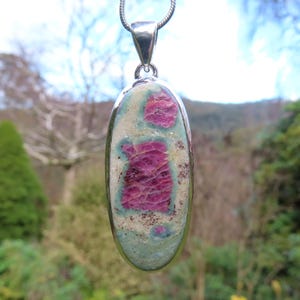 May include: Oval pendant with a silver-coloured bail and chain. The pendant features a large, central ruby-coloured stone with a light blue and cream-coloured matrix. The stone has a polished finish and is set in a silver-coloured bezel.