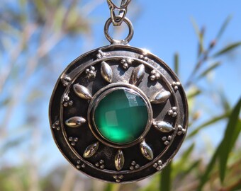 May include: A silver pendant necklace with a large, round, dark silver pendant. The pendant features a faceted, round, green gemstone in the center, surrounded by silver leaf-shaped accents and small silver beads. The necklace is displayed against a blurred green and blue background.