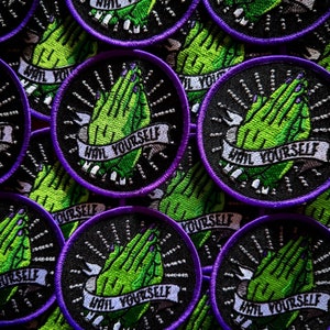 May include: Close-up of multiple black and purple embroidered patches with green hands in a praying position. The patches have the text "Hail Yourself" on a white banner.