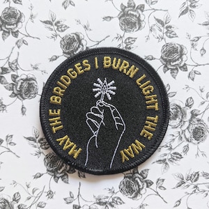 May include: A black embroidered patch with gold stitching. The patch features a hand holding a lit match and the text "May the bridges I burn light the way".