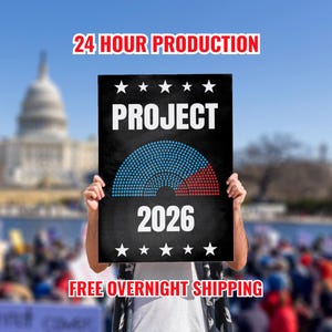 May include: A black sign with the word "PROJECT" in white capital letters, a semi-circle graphic divided into blue and red sections, and the year "2026". The sign also has white stars and the text "24 HOUR PRODUCTION".