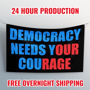 May include: A black banner with the words "DEMOCRACY NEEDS YOUR COURAGE" in blue and red block letters. The banner is suspended by a wire against a white wall. The text "24 HOUR PRODUCTION" and "FREE OVERNIGHT SHIPPING" are also displayed.