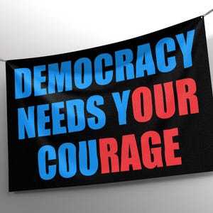 May include: A black banner with the words "DEMOCRACY NEEDS YOUR COURAGE" in blue and red block letters. The banner is hanging from a thin wire against a white background.