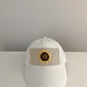 May include: White baseball cap with a beige patch featuring an embroidered sunflower in brown and yellow.