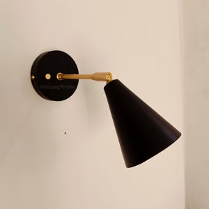Puede incluir: Una lámpara de pared negra con un brazo dorado y una pantalla cónica negra. La lámpara está montada en una pared blanca.