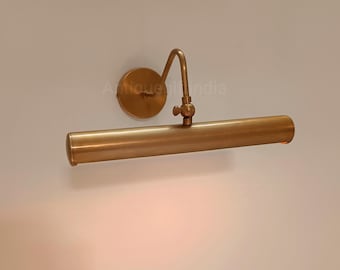 May include: A brass-toned wall-mounted picture light. The light has a cylindrical shade, a curved arm, and a round backplate. The fixture is designed to illuminate artwork or other wall-mounted items. The light is mounted on a white wall.