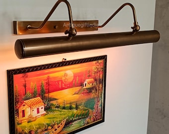 Op de afbeelding: Een messing schilderijlamp met twee verstelbare armen en een lange, cilindrische kap. De lamp is gemonteerd op een witte muur en belicht een ingelijst schilderij van een zonsonderganglandschap.