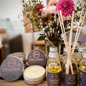 May include: A selection of reed diffusers and candles in glass bottles with black labels. The labels feature the brand name "Blystra" and the scents "Citronella & Lemongrass", "Lemongrass & Ginger", and "FairyDust". The bottles are arranged on a wooden surface with a bouquet of pink flowers in a glass vase.