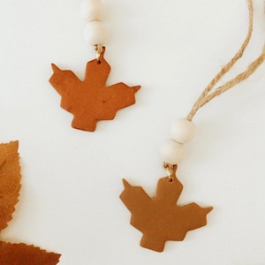 May include: Two brown clay maple leaf ornaments with gold accents hanging from twine with white wooden beads.