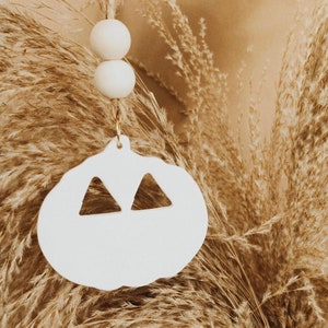May include: White ceramic pumpkin ornament with two triangle cutouts for eyes. The ornament is hanging from a string with two white wooden beads.