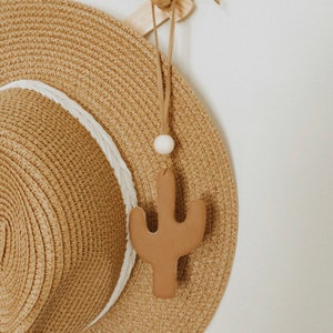 Peut inclure: Un chapeau de paille brun avec un bandeau blanc suspendu à un crochet en bois avec un cordon en cuir brun et un charme en céramique cactus brun.
