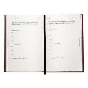May include: A brown leather-bound gratitude journal with a black ribbon bookmark. The journal has two pages open, each with a quote and prompts for reflection. The quote on the left page is "The trick is to be grateful when your mood is high and graceful when it is low." by Richard Carlson. The quote on the right page is "What seems insignificant when you have it is important when you need it." by Franz Grillparzer.